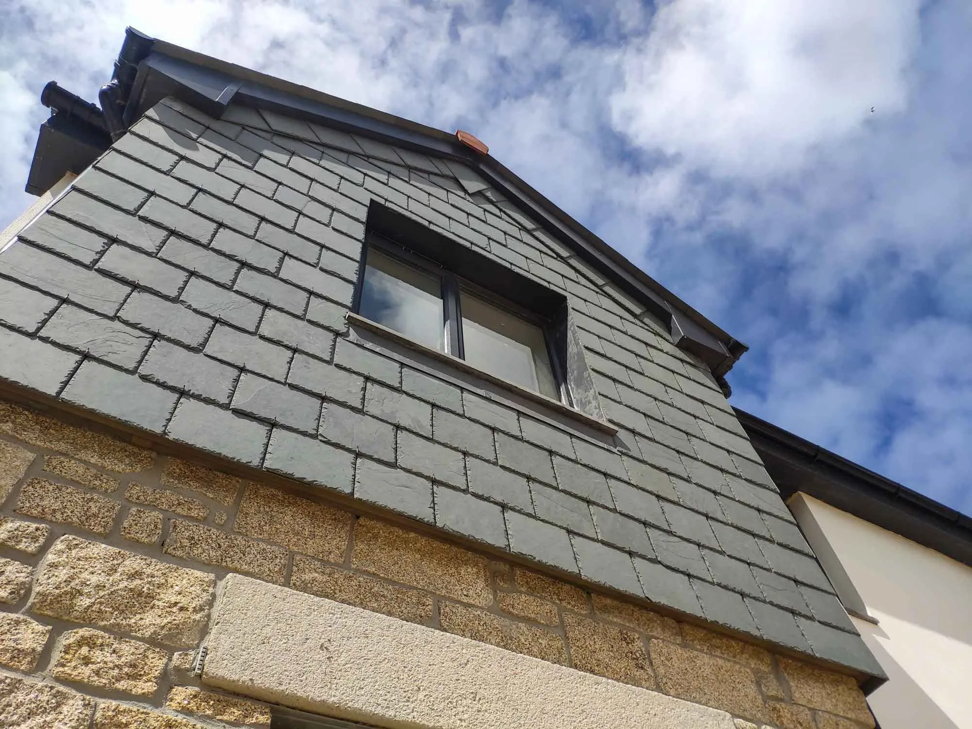 Dormer window clad with grey slate tiles on stone and plaster wall under blue sky